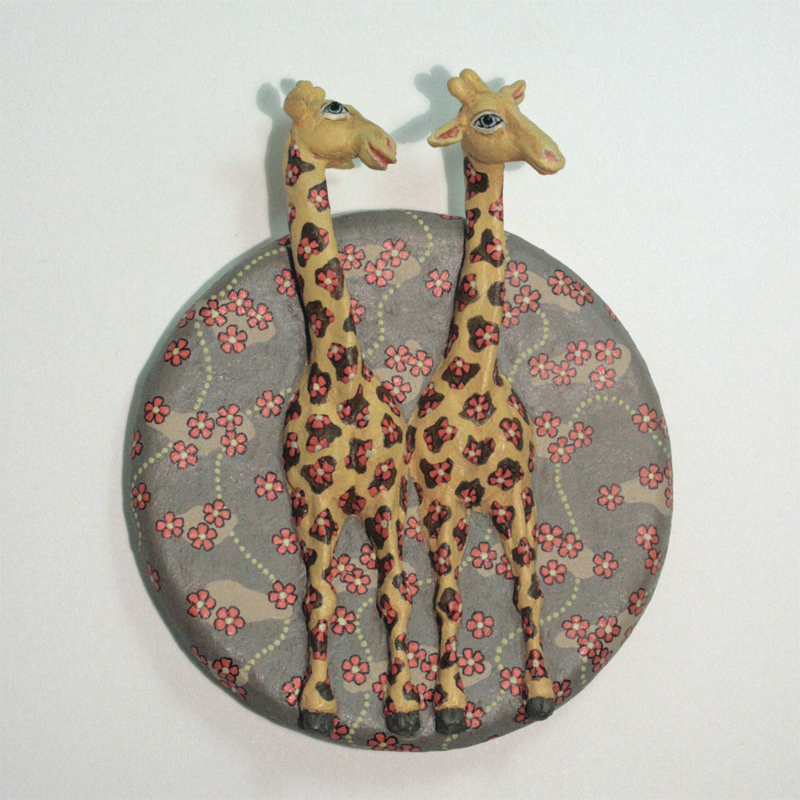 zwei gelbe Giraffen auf einer runden Platte zum Aufhängen, der Hintergrund ist hell braungrau, darauf rote Blüten, die auch bei den Giraffen auf den Flecken sitzen. Ansicht frontal.