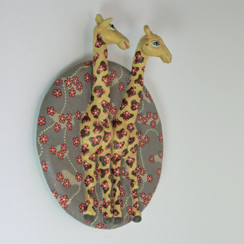 zwei gelbe Giraffen auf einer runden Platte zum Aufhängen, der Hintergrund ist hell braungrau, darauf rote Blüten, die auch bei den Giraffen auf den Flecken sitzen. Ansicht halb seitlich.