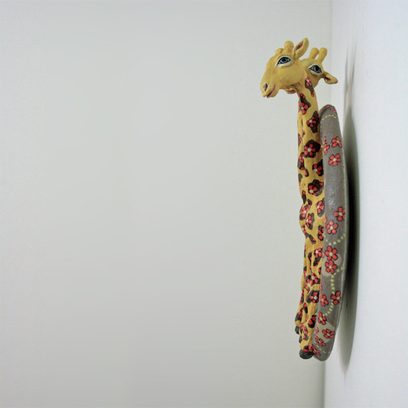 zwei gelbe Giraffen auf einer runden Platte zum Aufhängen, der Hintergrund ist hell braungrau, darauf rote Blüten, die auch bei den Giraffen auf den Flecken sitzen. Ansicht seitlich.