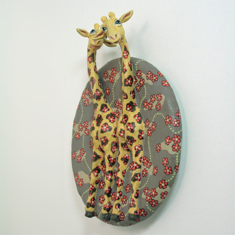 zwei gelbe Giraffen auf einer runden Platte zum Aufhängen, der Hintergrund ist hell braungrau, darauf rote Blüten, die auch bei den Giraffen auf den Flecken sitzen. Ansicht halb seitlich.