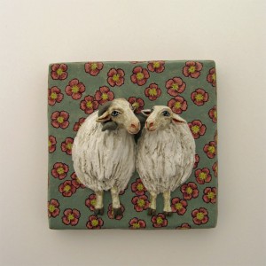 zwei Schafe, ein Widder und ein weibliches, die sich offenbar mögen, die Köpfe einander zugewandt. Auf matt graugrünem Grund mit rot-gelben Blüten.